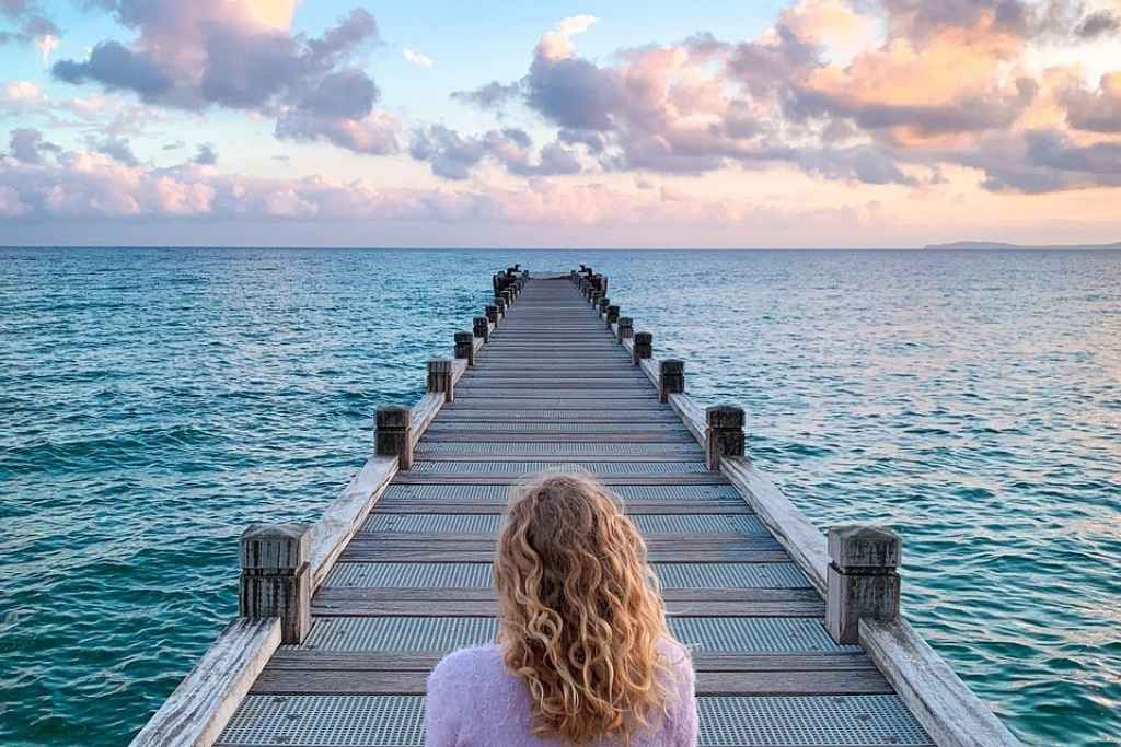 Counselling can help – woman seeking self-discovery Woman sitting alone on a long wooden pier, gazing into the distance in quiet contemplation, symbolising a journey of self-discovery and emotional healing through counselling.